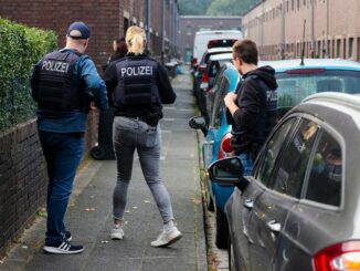 Beamte der Polizei stehen auf einer Straße vor einem Haus in Duisburg, das gerade durchsucht wird.