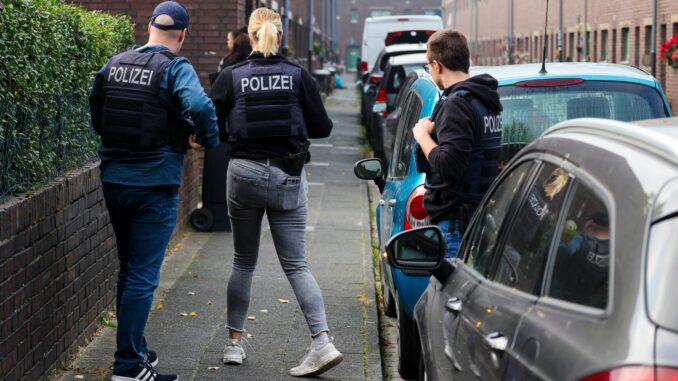 Beamte der Polizei stehen auf einer Straße vor einem Haus in Duisburg, das gerade durchsucht wird. 